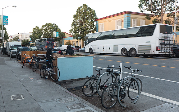 The stark demographics of San Francisco show up as two luxury buses dedicated to shuttling tech workers from SF to their workplaces in Silicon Valley cruise by a bicycle-filled parklet in front of the collectively-run bakery Arizmendi.